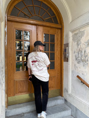 Person wearing a white t-shirt with red text and graphics, black pants, and a black cap standing in front of a wooden door.