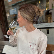 Woman sitting outdoors holding a glass of white wine, wearing a 'Cats & Wine' shirt.